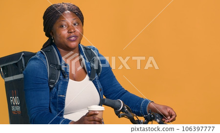 While delivering products young female courier on bicycle takes quiet break to enjoy cup of coffee. Delivery woman quenches her thirst with drink while offering prompt food delivery to neighborhood. 106397777