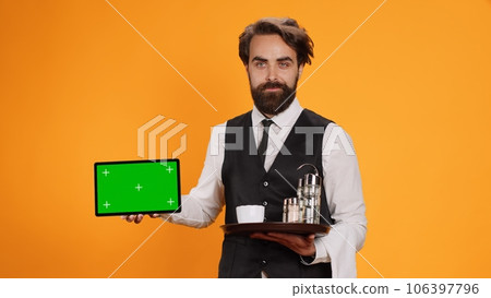 Smiling butler presents greenscreen on tablet, posing with confidence against yellow background. Young adult worker showing isolated template on smartphone display in front of camera. 106397796