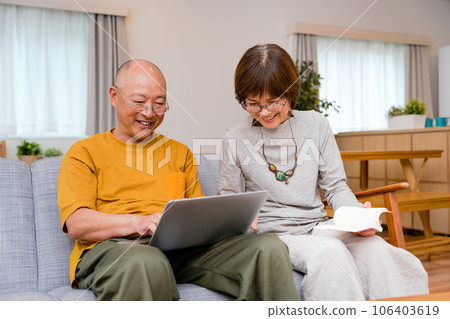 An elderly couple sitting on the sofa in the living room at home, a man operating a laptop on his lap and a woman reading a book. 106403619