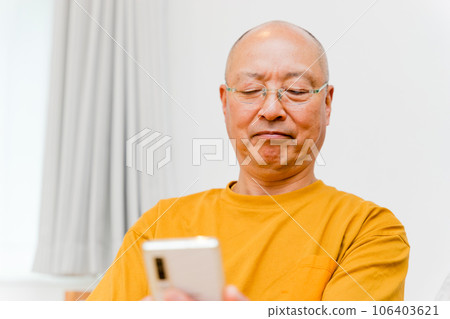 An elderly man wearing reading glasses operates his smartphone screen from a distance. An elderly man wearing reading glasses operates his smartphone screen from a distance. 106403621