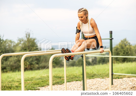 Sporty young female working out calisthenics in an outdoor gym using parallel bars. Beautiful blonde girl with braids works out outdoors doing push-ups on an elevated bar Sporty young female working out calisthenics in an outdoor gym using parallel bars. Beautiful blonde girl with braids works out outdoors doing push-ups on an elevated bar 106405602