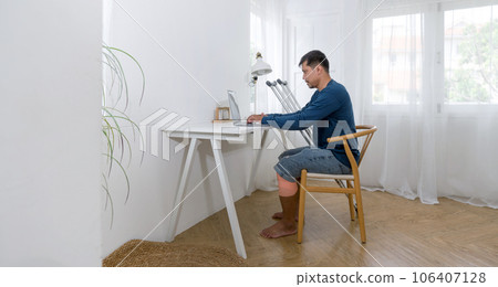 A man with a broken leg immersed in his work, sitting in an office or home-based workstation. He's engrossed in various tasks on his laptop, conveying a sense of productivity and focus. 106407128