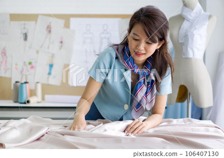 Young asian dressmaker in striped scarf spread out a piece of fine beige fabric on workbench preparing to create cloth pattern. Fashion designer room. Young asian dressmaker in striped scarf spread out a piece of fine beige fabric on workbench preparing to create cloth pattern. Fashion designer room. 106407130