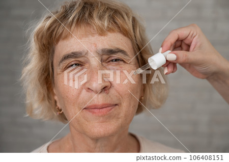 Close-up portrait of an old woman applying hyaluronic acid serum with a pipette. Anti-aging face care.  106408151
