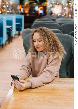 Young woman with curly hair is sitting at a cafe table and looking at her smartphone 106410794