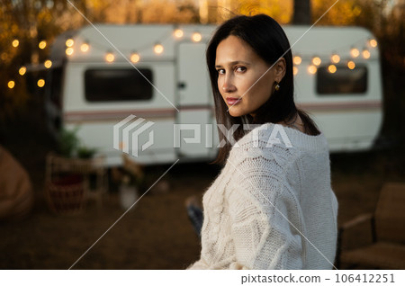 A pensive Caucasian woman in a white knitted sweater is resting in a warm autumn near a motorhome. Travel by camper.  106412251