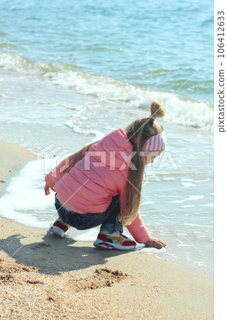 Little girl with long blond hair pink coat plays on the beach with sand shells stones looks at the sea waves sunny day. Little girl with long blond hair pink coat plays on the beach with sand shells stones looks at the sea waves sunny day. 106412633