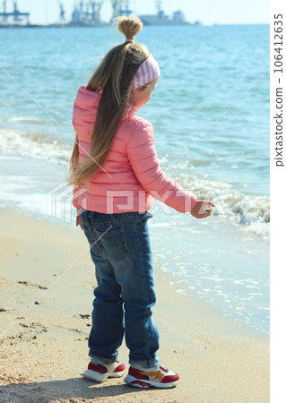 Little girl with long blond hair pink coat plays on the beach with sand shells stones looks at the sea waves sunny day. 106412635