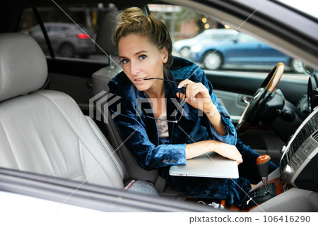 Beautiful blonde in blue suit looks at camera in car, holds laptop and holds glasses in her hand. 106416290