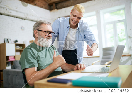 Cooperation of adults and youngsters. Young man helping senior businessman with technological issues on laptop Cooperation of adults and youngsters. Young man helping senior businessman with technological issues on laptop 106419280