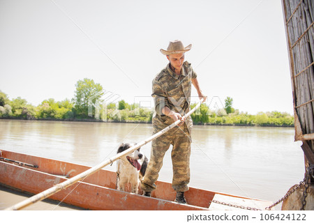 Active male fisher with dog sailing from dock in old boat, going fishing in sunny day. Front view of man in fatigues holding stick to push off from pier, while looking down. Concept of fishery. 106424122