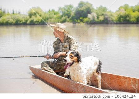 Focused young man in fatigues sailing along river with nice dog on board, while fishing in countryside. Side view of bored fisherman looking at float, while holding rod at daytime. Concept of fishing. 106424124