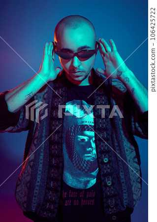 Studio shot of a young tattoed bald man posing against a colorful background. 90s style. Studio shot of a young tattoed bald man posing against a colorful background. 90s style. 106425722