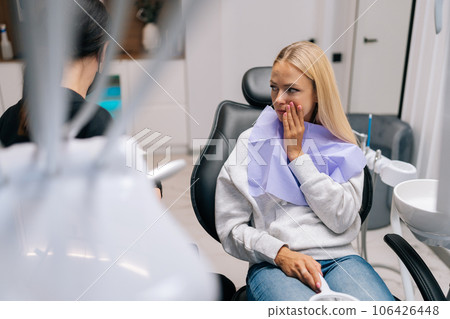 Side view of unrecognizable female dentist talking with blonde young woman patient complaining of toothache holding hand on cheek at dentist office. Concept of stomatology, dentistry and orthodontics. 106426448