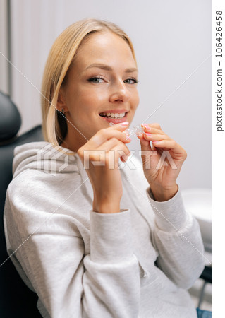 Vertical portrait of smiling blonde female patient holding invisible braces aligner sitting on chair in dentistry clinic. Close up of mobile orthodontic appliance for dental correction. Vertical portrait of smiling blonde female patient holding invisible braces aligner sitting on chair in dentistry clinic. Close up of mobile orthodontic appliance for dental correction. 106426458