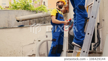 Competent mechanic holding electric hand drill stepping down from folding ladder after finishing servicing rooftop hvac system while colleague assembles manometer to check refrigerant levels Competent mechanic holding electric hand drill stepping down from folding ladder after finishing servicing rooftop hvac system while colleague assembles manometer to check refrigerant levels 106429144