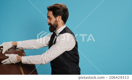 Friendly bellboy indicates satisfaction, showcasing role as professional hotel concierge employee in hospitality industry. Skilled doorman with white gloves gloves carrying suitcase in studio. 106429313