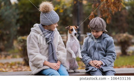 Brother and sister sit in an embrace with a dog on a bench for a walk in the autumn park. Boy, girl and jack russell terrier.  106433136