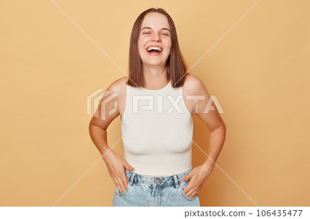 Laughing positive brown haired young woman wearing casual clothing standing isolated over beige background lookign at camera hearing funny joke expressing positive emotions Laughing positive brown haired young woman wearing casual clothing standing isolated over beige background lookign at camera hearing funny joke expressing positive emotions 106435477