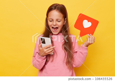 Clicking the like button on a blog post. Recommending viral content to followers. Amazed overjoyed little girl holding blogger heart like icon and smartphone isolated over yellow background 106435526