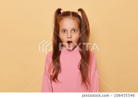 Shocked surprised little girl with ponytails wearing pink sweatshirt isolated over beige background looking at something astonished with widely open mouth and big eyes. Shocked surprised little girl with ponytails wearing pink sweatshirt isolated over beige background looking at something astonished with widely open mouth and big eyes. 106435546