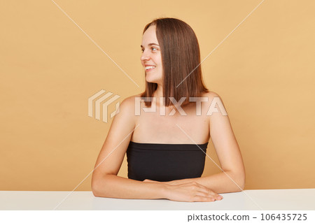 Cheerful smiling brown haired woman wearing black top posing isolated over beige background looking away at copy space for advertisement mockup for promotional text. Cheerful smiling brown haired woman wearing black top posing isolated over beige background looking away at copy space for advertisement mockup for promotional text. 106435725