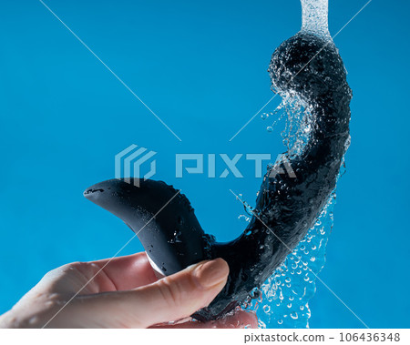 A woman washes a black prostate stimulator under running water on a blue background. Sex toy hygiene concept. 106436348