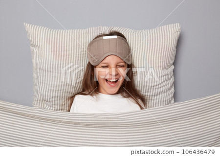 Happy little girl wearing white shirt and sleep eye mask lies under blanket in bedroom isolated on gray background laughing out loud having fun in morning Happy little girl wearing white shirt and sleep eye mask lies under blanket in bedroom isolated on gray background laughing out loud having fun in morning 106436479