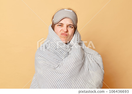 Displeased unhappy woman wrapped blanket and wearing sleeping eye mask isolated over beige background does not want to wake up early grimacing frowning face. 106436693