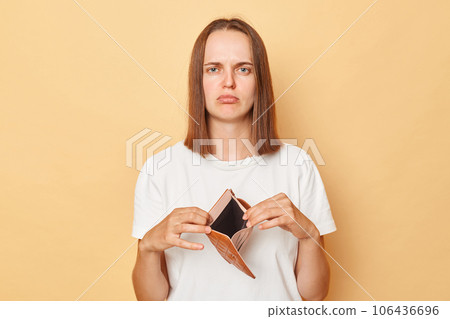 Sad upset young caucasian woman wearing white T-shirt showing empty wallet over isolated beige background has no money to spend looking at camera with displeased expression. 106436696