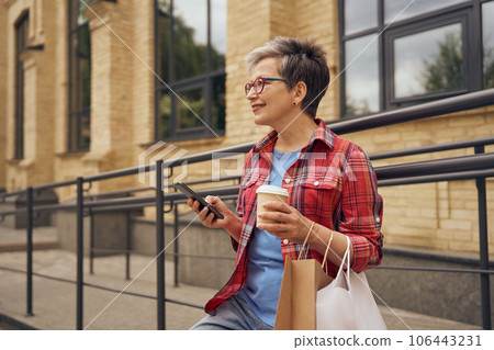 Stylish mature woman walking with her shopping bags, drinking coffee and use phone in city street Stylish mature woman walking with her shopping bags, drinking coffee and use phone in city street 106443231