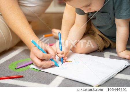 A beautiful little toddler boy of two or three years old with dad draws with markers in the album in the children's room at home sitting on the floor. Spending time with children. Selective focus 106444591