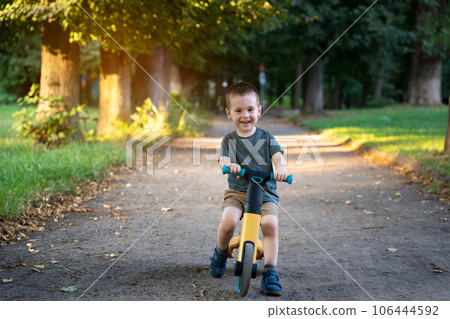 A happy smiling toddler boy of two or three years old rides a bicycle or balance bike in a city park on a sunny summer day. Toddlerhood and childhood concept. Selective focus A happy smiling toddler boy of two or three years old rides a bicycle or balance bike in a city park on a sunny summer day. Toddlerhood and childhood concept. Selective focus 106444592