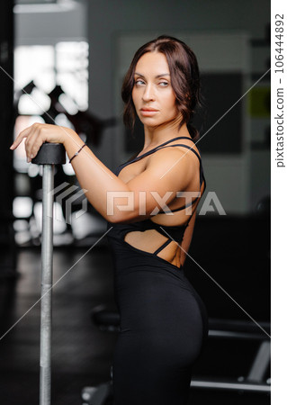 Female bodybuilder with long hair posing in gym with sports equipment. Strong woman with muscular body leaning on barbells in gym in dark atmosphere, looking at camera. Female bodybuilder with long hair posing in gym with sports equipment. Strong woman with muscular body leaning on barbells in gym in dark atmosphere, looking at camera. 106444892