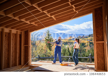 Father, mother and son building wooden frame house. Male designer in blue overalls showing future home, standing on terrace on construction site. Carpentry, family concept. Father, mother and son building wooden frame house. Male designer in blue overalls showing future home, standing on terrace on construction site. Carpentry, family concept. 106448653
