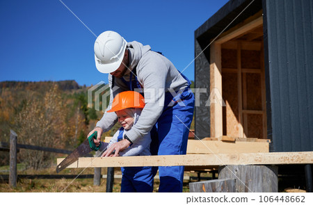 Father with toddler son building wooden frame house in the Scandinavian style barnhouse. Male builders using hand saw to cut boards on construction site on sunny day. Carpentry and family concept. 106448662