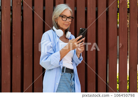 mature woman with gray hair and glasses listens to her favorite music in headphones while sitting on mature woman with gray hair and glasses listens to her favorite music in headphones while sitting on 106450798
