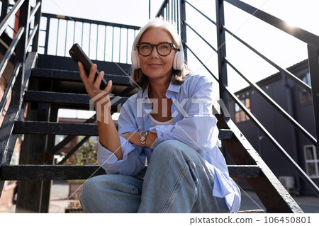 mature gray-haired woman in summer outfit listens to music with headphones while walking around the mature gray-haired woman in summer outfit listens to music with headphones while walking around the 106450801