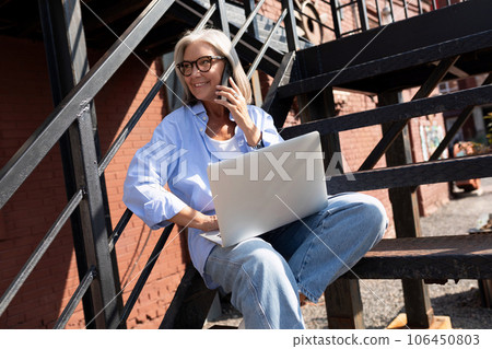 beautiful mature old woman with gray hair dressed makes a phone call while sitting on the street 106450803