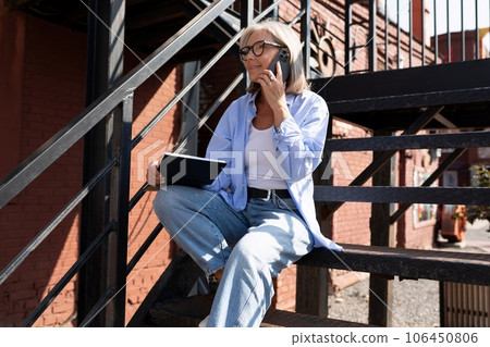 a well-groomed middle-aged woman with a bob haircut speaks on the phone while sitting with a laptop 106450806