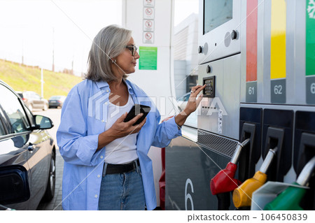 a mature woman with gray hair stands near a gas station and holds a smartphone 106450839