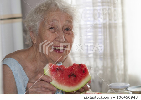 Smiling old senior woman holding a slice of fresh watermelon looking at camera at home background - healthy eating lifestyle concept 106450948