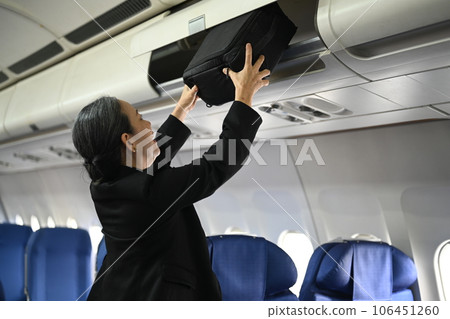 Smiling middle age woman passengers putting her luggage into the overhead compartment on an airplane Smiling middle age woman passengers putting her luggage into the overhead compartment on an airplane 106451260