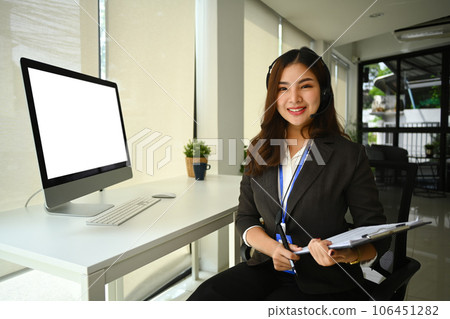 Portrait of call center telemarketer woman with headset sitting in front of blank computer monitor and smiling at camera Portrait of call center telemarketer woman with headset sitting in front of blank computer monitor and smiling at camera 106451282
