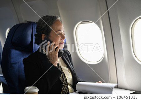 Positive middle aged businesswoman talking on mobile phone and looking away on the airplane during flight 106451315