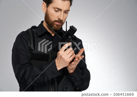 Focused dark-haired officer reloading weapon, getting ready to shoot indoors. Portrait of male cop looking at handgun, while recharging it, on gray studio background. Profession, weapon concept. 106451669