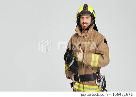 Happy caucasian firefighter in uniform showing thumb up in studio. Front view of confident bearded fireman giving like, while smiling to camera, on gray background. Concept of work, hand gesture. 106451671