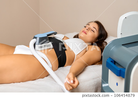 A youthful brunette client enjoys her body sculpting session, comfortably resting on a couch in the beauty salon. A youthful brunette client enjoys her body sculpting session, comfortably resting on a couch in the beauty salon. 106454879
