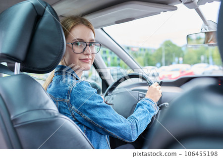 Teenage girl driver in glasses sitting behind wheel of car, looking at camera 106455198