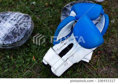 Shabby blue boxing gloves with black paws for practicing punches lie on the grass for street martial arts training Shabby blue boxing gloves with black paws for practicing punches lie on the grass for street martial arts training 106455317